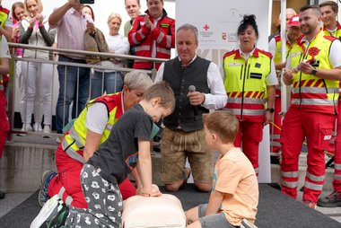 Ein Kind reanimiert eine Wiederbelebungspuppe, daneben ein weiteres Kind, im Hintergrund Sanitäter:innen, Zuschauer:innen und der Initiator des Weltrekordversuchs Dr. Ernst Toferer