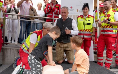 Ein Kind reanimiert eine Wiederbelebungspuppe, daneben ein weiteres Kind, im Hintergrund Sanitäter:innen, Zuschauer:innen und der Initiator des Weltrekordversuchs Dr. Ernst Toferer