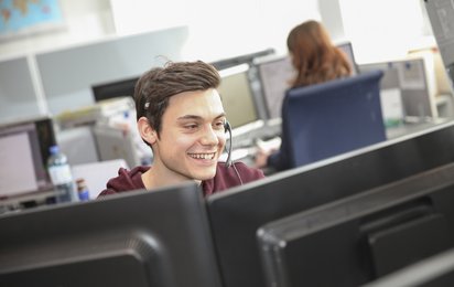 Ein Mitarbeiter des Service-Centers der Blutspendezentrale sitzt mit Headset vor zwei Monitoren und lächelt