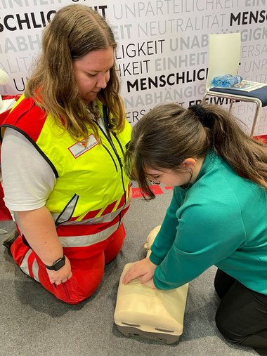 Eine Frau übt knieend an einer Trainingspuppe die Herzmassage und wird dabei von eines ebenfalls knieenden Sanitäterin beobachtet.
