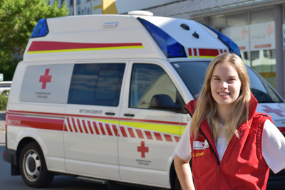 Sanitäterin Anna-Maria steht vor einem Rettungswagen und trägt die markante rote Uniformjacke.
