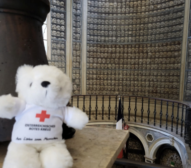 Erklärbär Bärli blickt in die Kamera und befindet sich dabei in der Knochenkapelle in Solferino. In dieser Kapelle werden die Schädel von 1413 Gefallenen der Schlacht und Knochen von ca. 7000 weiteren Opfern aufbewahrt