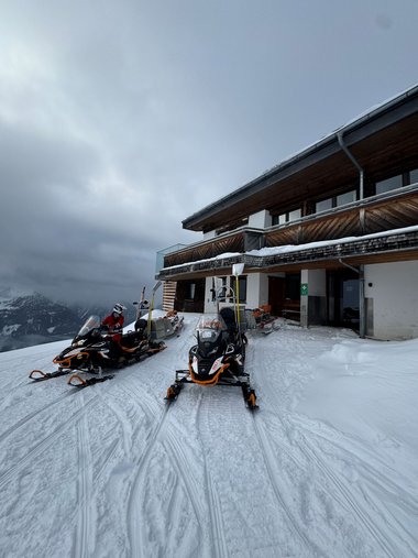 Zwei Skidoos stehen vor einem Gebäude im Schnee zum Einsatz bereit, der Himmel ist bewölkt.