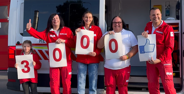 Anlässlich des 3000sten Followers auf Facebook steht eine Gruppe von Rotkreuz-Mitarbeitern mit der Zahl 3000 auf Zetteln vor einem Rotkreuz-Auto.
