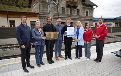 Acht Personen stehen an einem Bahnsteig und blicken in die Kamera. Eine Person hält eine Tafel in der Hand, eine andere ein Zertifikat.