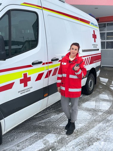 Eine Frau steht mit Rotkreuzjacke vor einem Rettungswagen und hält ein Funkgerät in der Hand