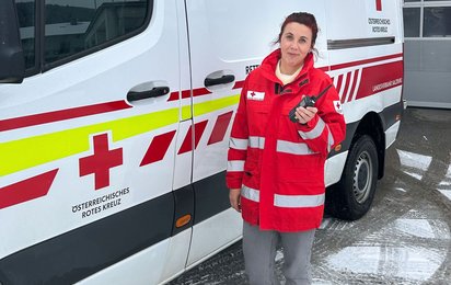Eine Frau steht mit Rotkreuzjacke vor einem Rettungswagen und hält ein Funkgerät in der Hand