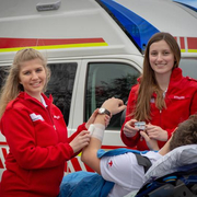 Carina und Helena sind freiwillige Sanitäterinnen - © OÖRK Zwei weibliche Sanitäterinnen vor einem Rettungsauto wie sie einen Patienten betreuen.