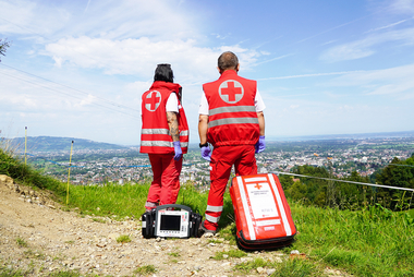 Zwei Sanitäter:innen blicken vom Karren aus in die Talschaft. Sie haben einen Notfallrucksack und einen Defibrillator dabei.