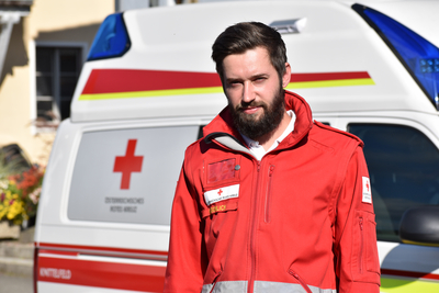 Sanitäter Christian steht vor einem Rettungswagen und trägt die markante rote Uniformjacke.