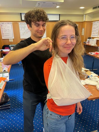 Ein Mann legt einer Frau einen Verband am Arm an und befestigt ihn hinter ihrem Kopf.