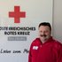 Martin Wenzel mit roter Jacke vor einer Wand mit einem Schild mit dem Logo des Roten Kreuzes Salzburg, darunter der Slogan "Aus Liebe zum Menschen".