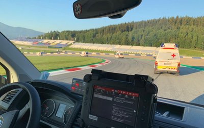 Einige Rettungswagen befinden sich auf einer Rennstrecke (dem Red Bull Ring) und beziehen grade ihre zugewiesenen Posten. Man sieht eine Fahrerkabine von Innen und weitere Rettungswagen vor dem Fahrzeug.