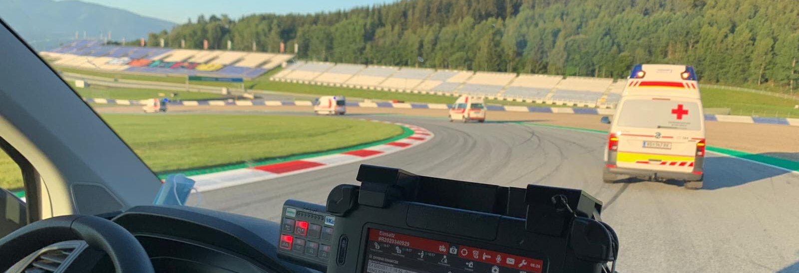 Einige Rettungswagen befinden sich auf einer Rennstrecke (dem Red Bull Ring) und beziehen grade ihre zugewiesenen Posten. Man sieht eine Fahrerkabine von Innen und weitere Rettungswagen vor dem Fahrzeug.
