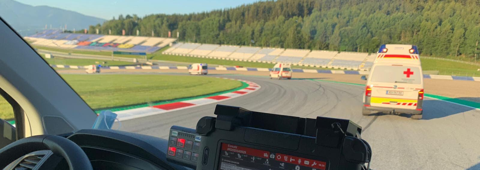 Einige Rettungswagen befinden sich auf einer Rennstrecke (dem Red Bull Ring) und beziehen grade ihre zugewiesenen Posten. Man sieht eine Fahrerkabine von Innen und weitere Rettungswagen vor dem Fahrzeug.