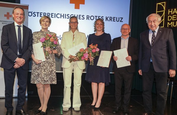 Sechs Personen stehen bei einer Veranstaltung des Österreichischen Roten Kreuzes auf einer Bühne. Drei Frauen und drei Männer halten Urkunden und Blumensträuße in den Händen.