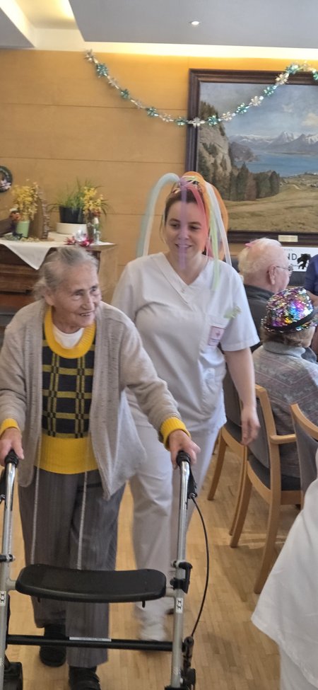 Eine Bewohnerin geht mit ihrem Rollator, begleitet wird sie von einer Pflegemitarbeiterin, die einen Hut aus bunten Luftballonen trägt.