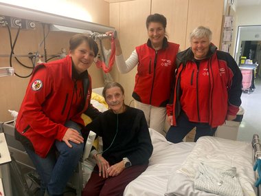 Eine Dame, Margarete, sitzt auf einem Krankenbett. Um sie herum stehen drei Damen, die sie auf ihrer Fahrt nach Villach begleitet haben.