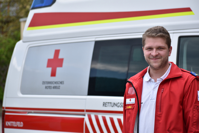 Sanitäter Daniel steht vor einem Rettungswagen und trägt die markante rote Uniformjacke.