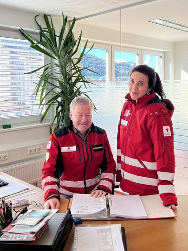Ein Mann sitzt in Rotkreuzjacke an einem Schreibtisch, rechts von ihm steht eine Frau in Rotkreuzjacke.