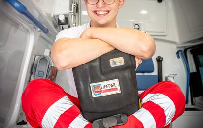 Zivildiener Alexander sitzt auf der Trage im Rettungsauto und hält einen Defibrillator in der Hand.