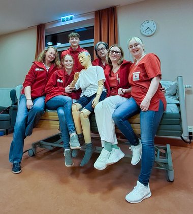 Eine Gruppe von Personen in Rotkreuz-Kleidung sitzt zusammen auf einem Bett und hält eine Simulations-Puppe.