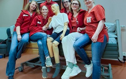 Eine Gruppe von Personen in Rotkreuz-Kleidung sitzt zusammen auf einem Bett und hält eine Simulations-Puppe.