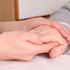 Die Hände einer jüngeren Frau umschließen eine Hand eines alten Menschen mit goldenem Ring und grauem Pulloverärmel.
