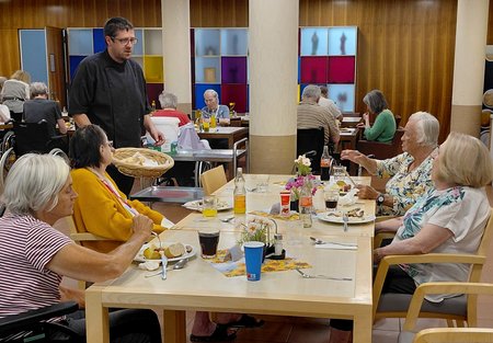 Ein Koch steht mit einem Brotkorb vor einem Esstisch, an dem vier Bewohnerinnen des Seniorenwohnhauses sitzen