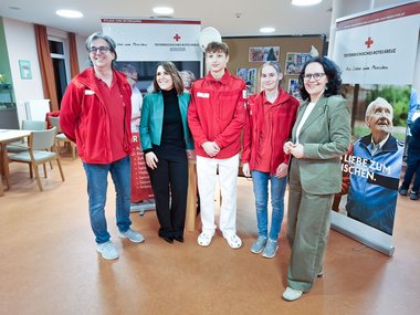 Fünf Personen in roten Jacken des Österreichischen Roten Kreuzes sowie zwei ältere Personen stehen zusammen in einem Büroraum.