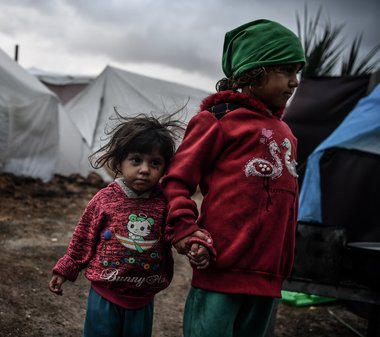 © ICRC / Abed Zaqout Zwei kleine Kinder stehen Hand in Hand auf schlammigen Boden vor Zelten in Gaza.
