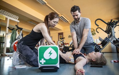 Notfall im Fitnesscenter, Helferin und Helfer verwenden Defibrillator bei Patientin, Querformat