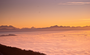 Man sieht einen wunderschönen Sonnenuntergang über einer Wolkendecke und Bergen im Hintergrund.