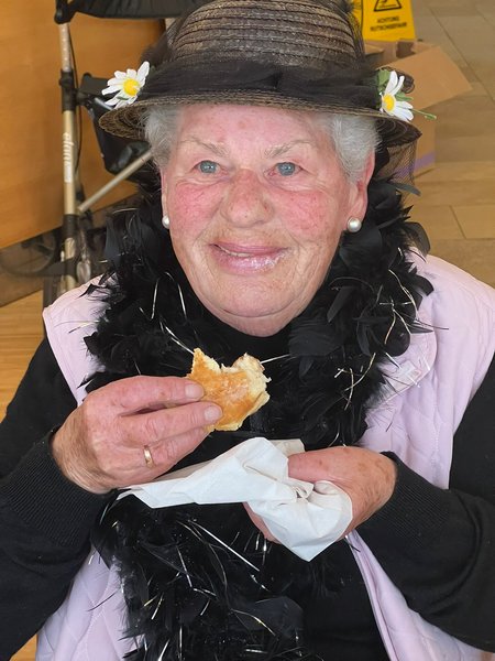 Eine mit Hut und Federboa verkleidete Bewohnerin genießt einen Krapfen. Auf ihren Lippen sieht man Spuren des Staubzuckers vom Krapfen.