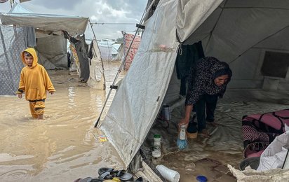 Kind geht durch Zeltstadt bei heftigem Regen - eine Frau versucht ihr Zelt trocken zu halten.