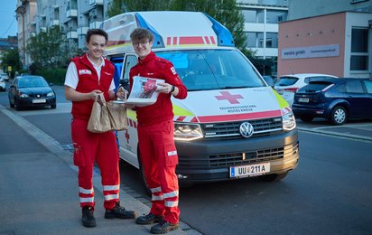 Versorgung Linz Marathon - © Credit: OÖRK/ Werner Asanger Zwei Rettungssanitäter stehen vor ihrem Auto und halten beide ein toll gefülltes Versorgungssackerl hoch mit Essen für den Tag