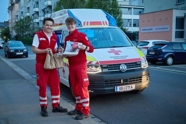 Zwei Rettungssanitäter stehen vor ihrem Auto und halten beide ein toll gefülltes Versorgungssackerl hoch mit Essen für den Tag