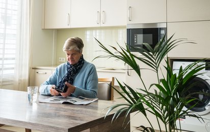 Eine ältere Dame sitzt am Tisch in ihrer Wohnung und scrollt auf ihrem Smartphone.