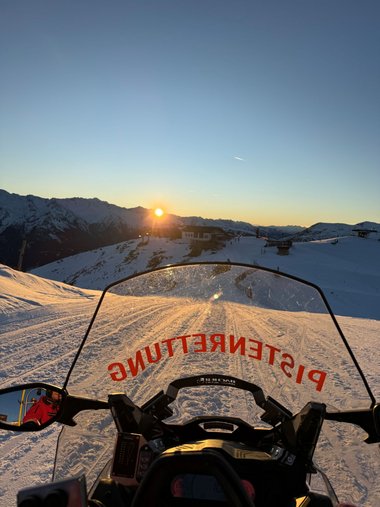 Aufnahme aus der Fahrposition auf einem Skidoo mit Blick in das Skigebiet. Im Hintergrund geht über den Bergen die Sonne unter.