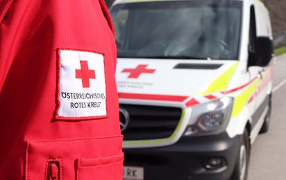 Ein im Anschnitt gezeigter Arm in Rotkreuz-Jacke, dahinter leicht verdeckt ein Rettungswagen.