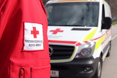 Ein im Anschnitt gezeigter Arm in Rotkreuz-Jacke, dahinter leicht verdeckt ein Rettungswagen.