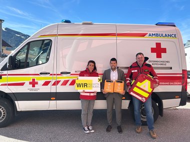 Drei Personen stehen vor einem Rettungswagen. Die Dame links trägt eine Rotkreuz-Jacke und hält ein Raiffeisen-Schild in der Hand. Der Herr in der Mitte trägt Anzug und hält zwei Defibrillatoren. Der Herr rechts trägt ebenfalls eine Rotkreuz-Jacke und hält einen Notfall-Rucksack.