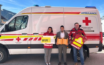 Drei Personen stehen vor einem Rettungswagen. Die Dame links trägt eine Rotkreuz-Jacke und hält ein Raiffeisen-Schild in der Hand. Der Herr in der Mitte trägt Anzug und hält zwei Defibrillatoren. Der Herr rechts trägt ebenfalls eine Rotkreuz-Jacke und hält einen Notfall-Rucksack.
