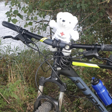 Bärli sitzt auf einem Fahrrad und blickt in die Kamera. Im Hintergrund sind Bäume und ein Fluss zu sehen.