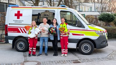Gruppenfoto vor einem Rettungswagen. Links eine Sanitäterin mit einem Blumenstrauß und einem großen, weißen Teddybären in der Hand. Rechts neben ihr ein Mann, der ein Baby im Arm hält. Neben ihm eine Frau mit Blumenstrauß und ganz rechts ein Sanitäter mit einem Blumenstrauß.