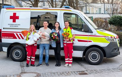 Gruppenfoto vor einem Rettungswagen. Links eine Sanitäterin mit einem Blumenstrauß und einem großen, weißen Teddybären in der Hand. Rechts neben ihr ein Mann, der ein Baby im Arm hält. Neben ihm eine Frau mit Blumenstrauß und ganz rechts ein Sanitäter mit einem Blumenstrauß.