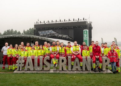 Ein Team an Sanitäter:innen steht hinter einem Schriftzug "#AustrianGP". Sie tragen Rotkreuz-Uniform und sind im Zuge des MotoGP-Rennens am Red Bull Ring im Einsatz.