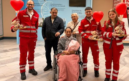 Rotkreuz-Mitarbeiter:innen mit der Patient:innen im Rollstuhl, sie hält einen Blumenstrauß in der Hand.