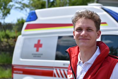 Sanitäter Gabriel steht vor einem Rettungswagen und trägt die markante rote Uniformjacke.