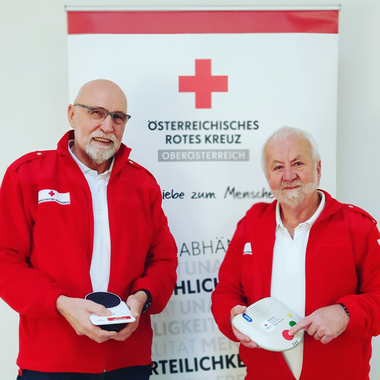 Rufhilfemonteure Charly und Karl stehen mit einem Rufhilfegerät vor dem Rotkreuz-Rollup